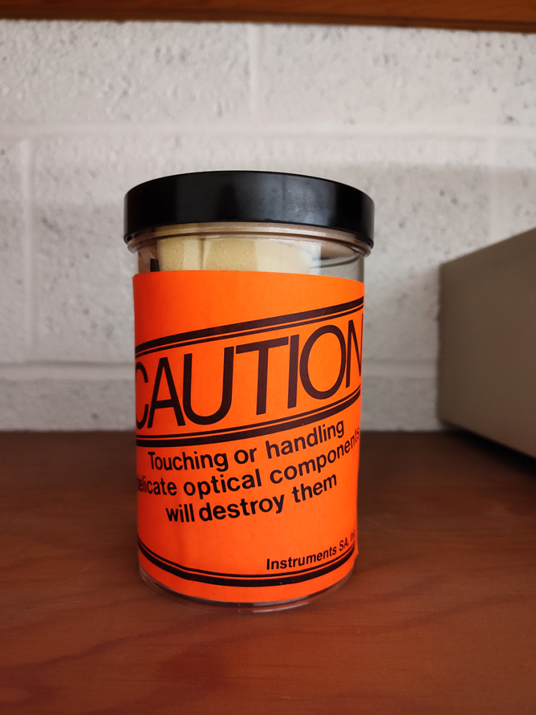 Orange warning label reads "CAUTION Touching or handling delicate optical components will destroy them"